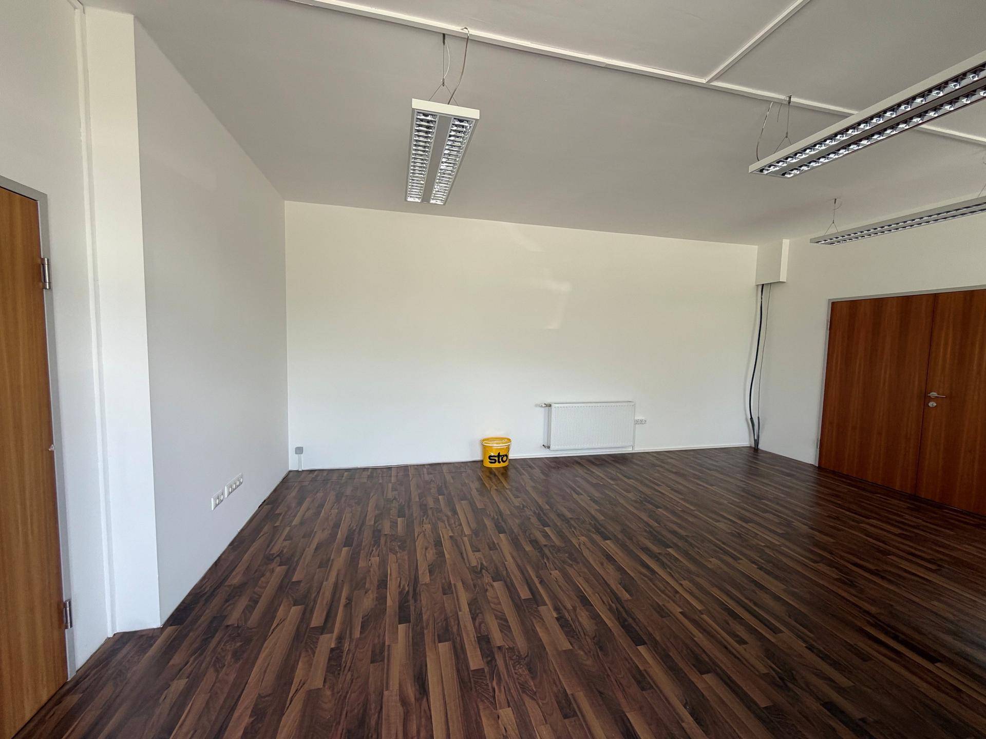 Leerer Gewerberaum mit dunklem Laminatboden, weißen Wänden und Deckenleuchten.