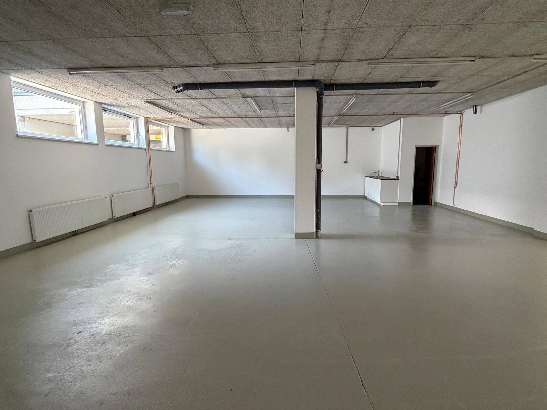 Leerer Gewerberaum mit Betonboden, weißen Wänden und sichtbaren Rohren an der Decke.