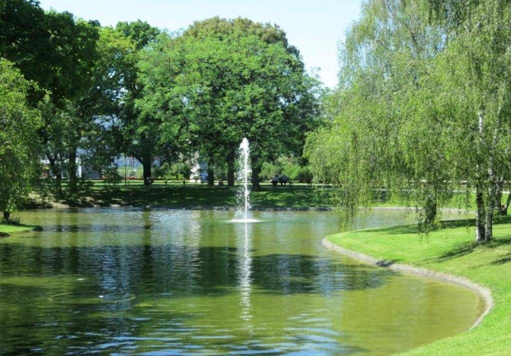 Idyllischer Teich mit Springbrunnen, umgeben von üppigem Grün, Bäumen und Rasenflächen.