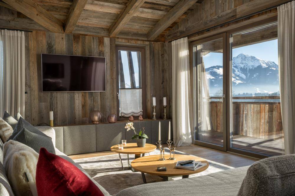 Lichtdurchflutetes Wohnzimmer mit Holzwänden, großem Fernseher und Panoramablick auf die Berge.