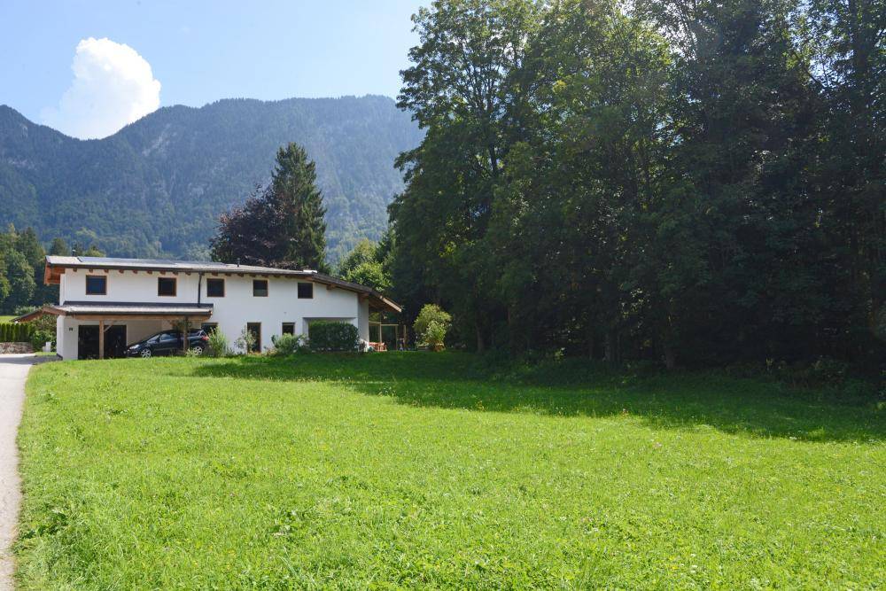 Modernes Wohnhaus mit Carport, umgeben von grünen Wiesen und majestätischen Bergen im Hintergrund.