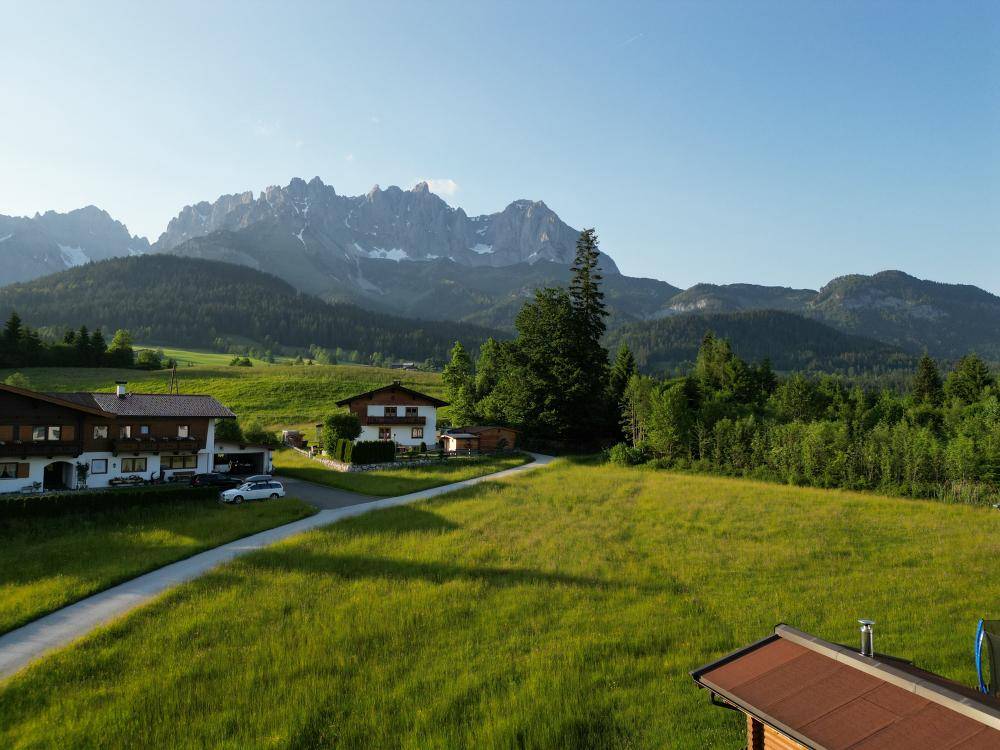 Panoramablick auf grüne Wiesen, eine Zufahrt und traditionelle Häuser vor einer imposanten Bergkette.