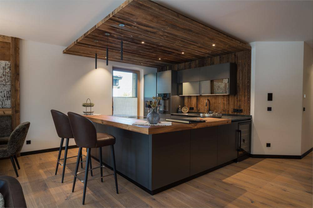 Moderne Küche mit dunklen Fronten, Holzdecke und praktischer Kücheninsel mit Barhockern.