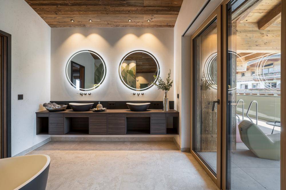 Elegantes Badezimmer mit zwei beleuchteten Spiegeln, Doppelwaschbecken und Balkonzugang.