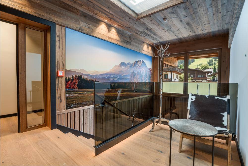 Heller Flur mit Holztreppe, Glasgeländer und einer beeindruckenden Berglandschaft als Wandbild.