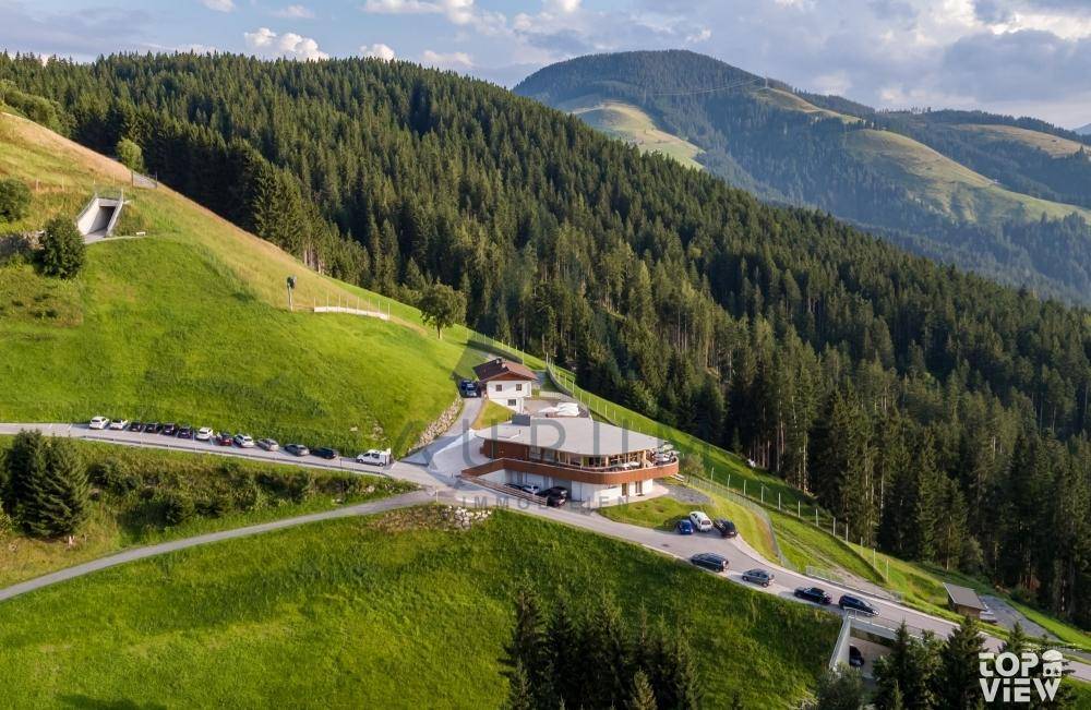 Luftaufnahme des Gebäudekomplexes und der Zufahrtsstraße, eingebettet in Wälder und Berge.