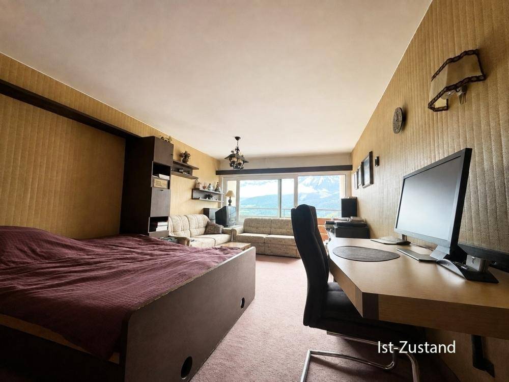 Wohnbereich im Ist-Zustand mit Bett, Schreibtisch und Sofa, bietet Bergblick durch das Fenster.