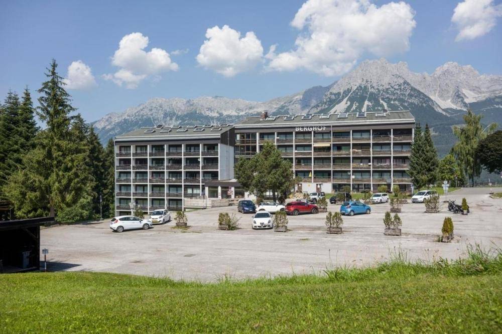 Großes Gebäude mit Parkplatz davor und majestätischen Bergen im Hintergrund unter blauem Himmel.