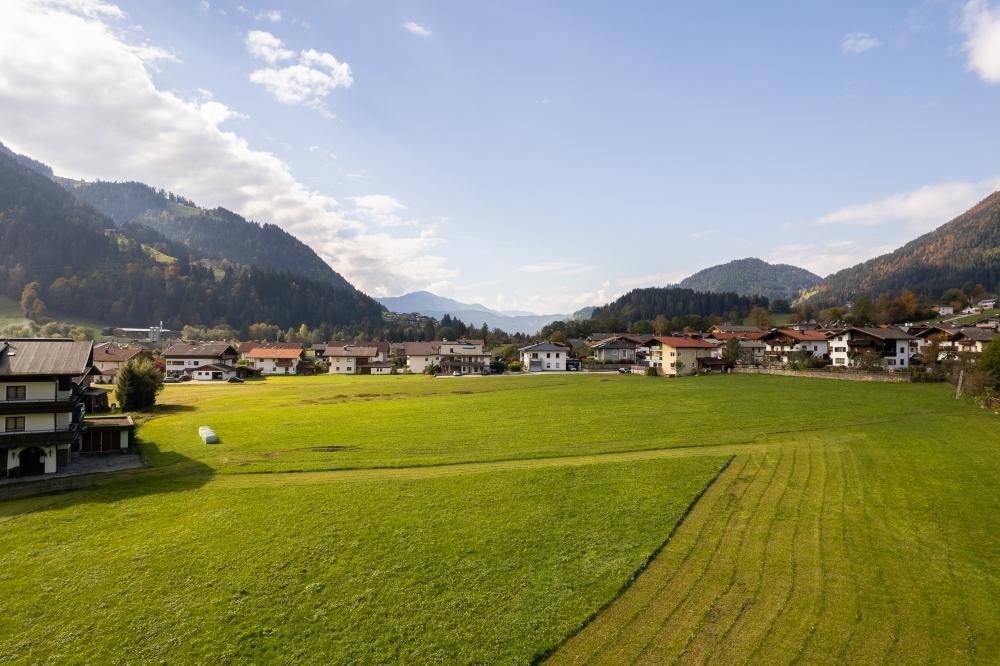 Weitläufige grüne Wiesen vor einer Reihe von Häusern in einem Dorf, mit bewaldeten Bergen am Horizont.