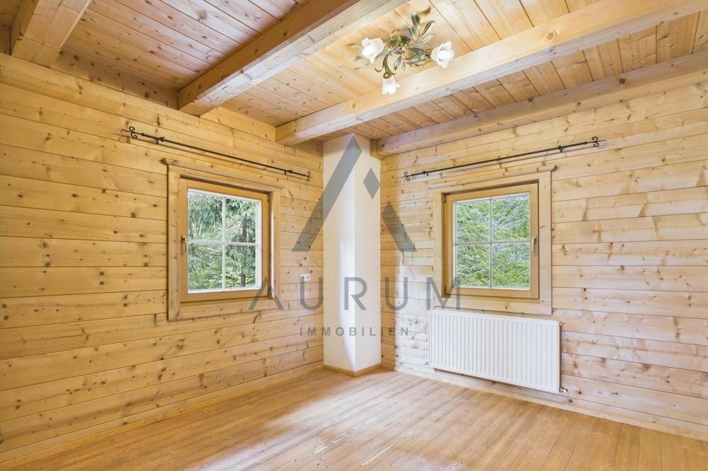Rustikales Zimmer mit Holzwänden, Holzbalkendecke und Fenstern mit Blick auf Berge und Bäume.