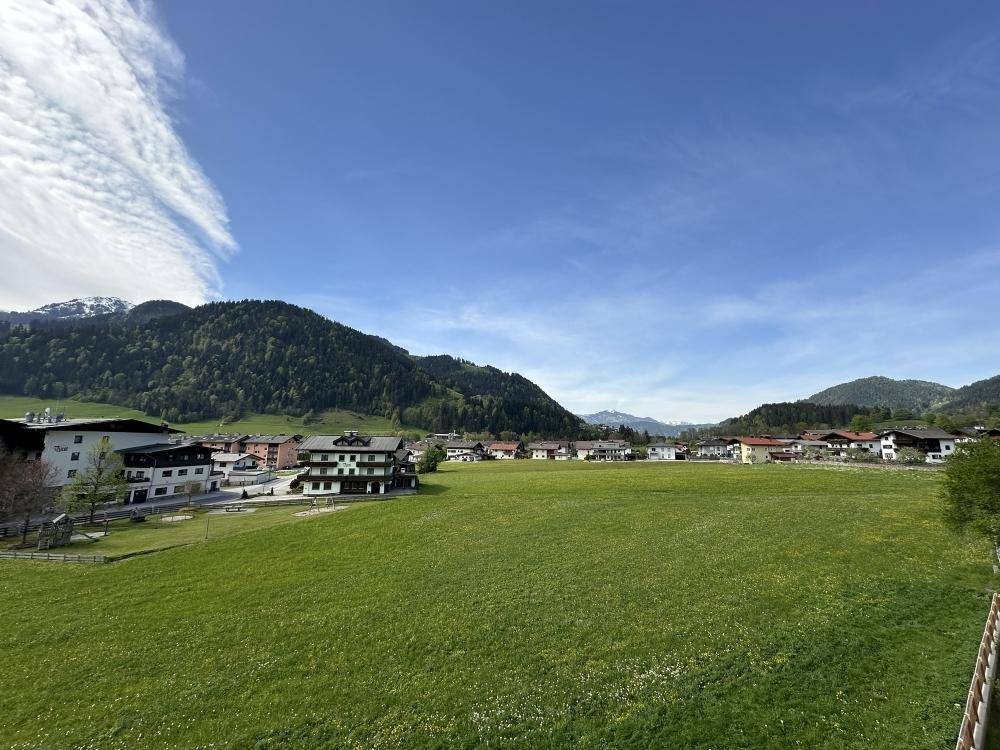 Weitläufige grüne Wiesen vor einer Kulisse aus Häusern und bewaldeten Bergen unter blauem Himmel.