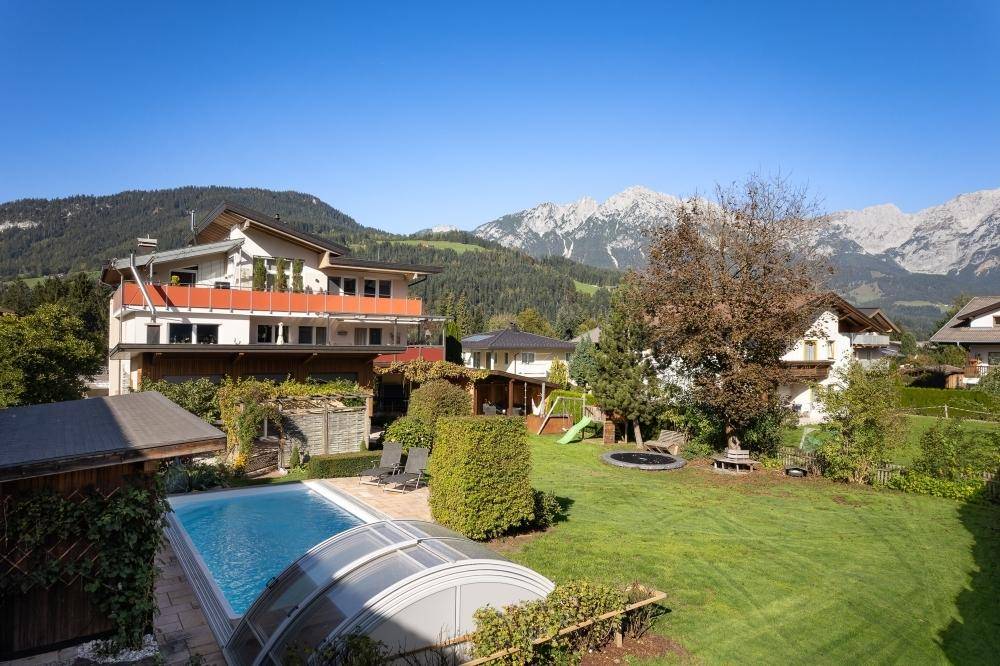 Weitläufiger Gartenbereich mit Pool, Spielplatz und Trampolin, umgeben von Hecken und mit Bergblick.