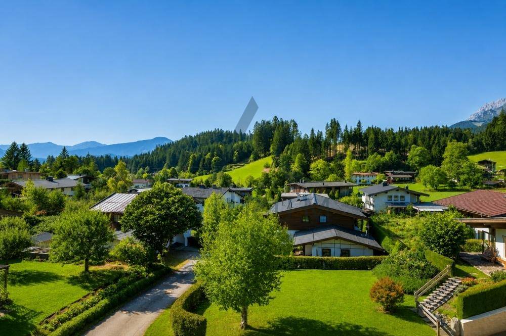 Weitläufiger Blick über eine ländliche Siedlung mit Häusern, umgeben von Wäldern und Bergen.