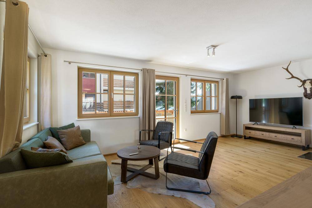 Gemütliches Wohnzimmer mit bequemer Couch, Holzboden und Blick ins Grüne durch die Fenster.