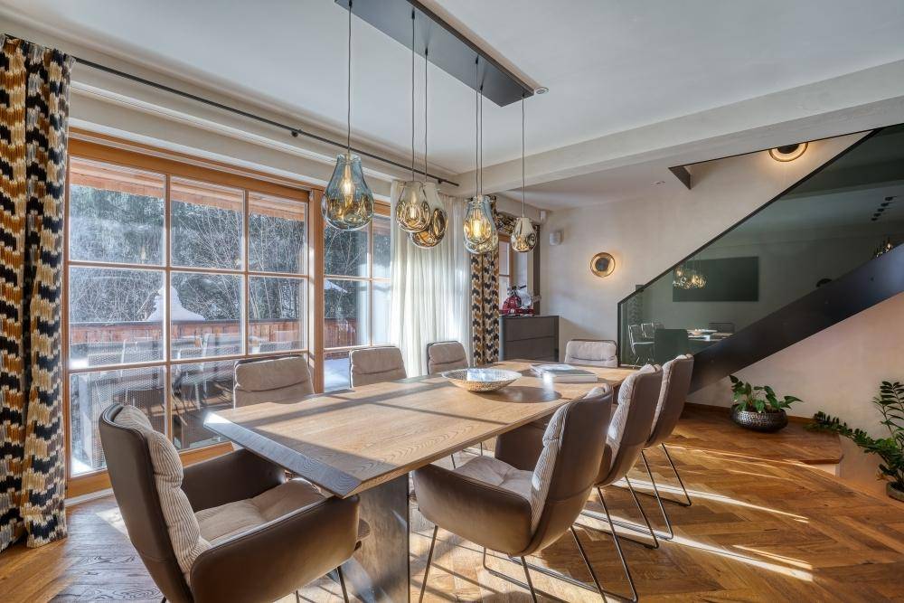 Eleganter Essbereich mit großem Holztisch, bequemen Stühlen und großen Fenstern mit Blick ins Grüne.