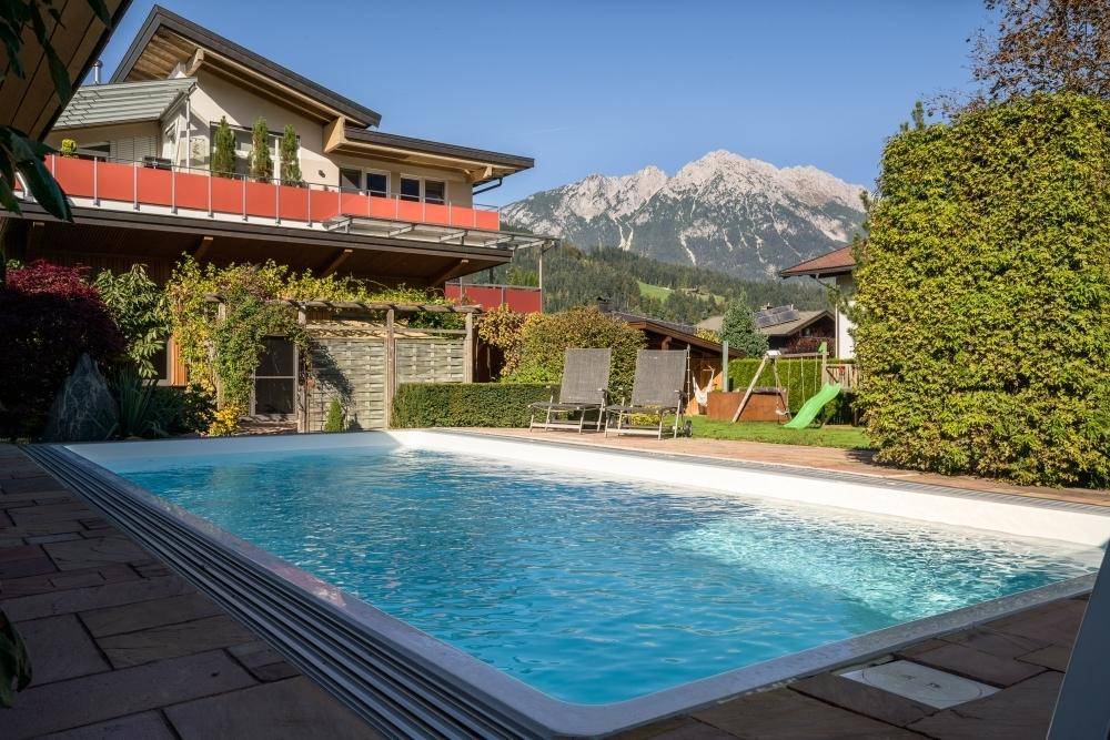 Großer Außenpool im Garten mit Liegestühlen, umgeben von Grünflächen und einem Spielplatz mit Bergblick.