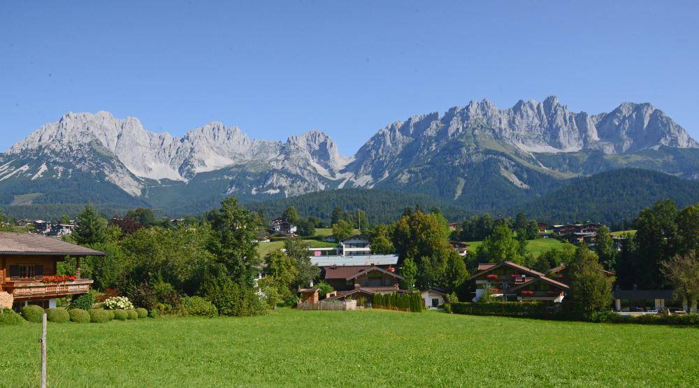 Weitläufige Landschaft mit mehreren Häusern, grünen Wiesen und beeindruckendem Alpenpanorama.