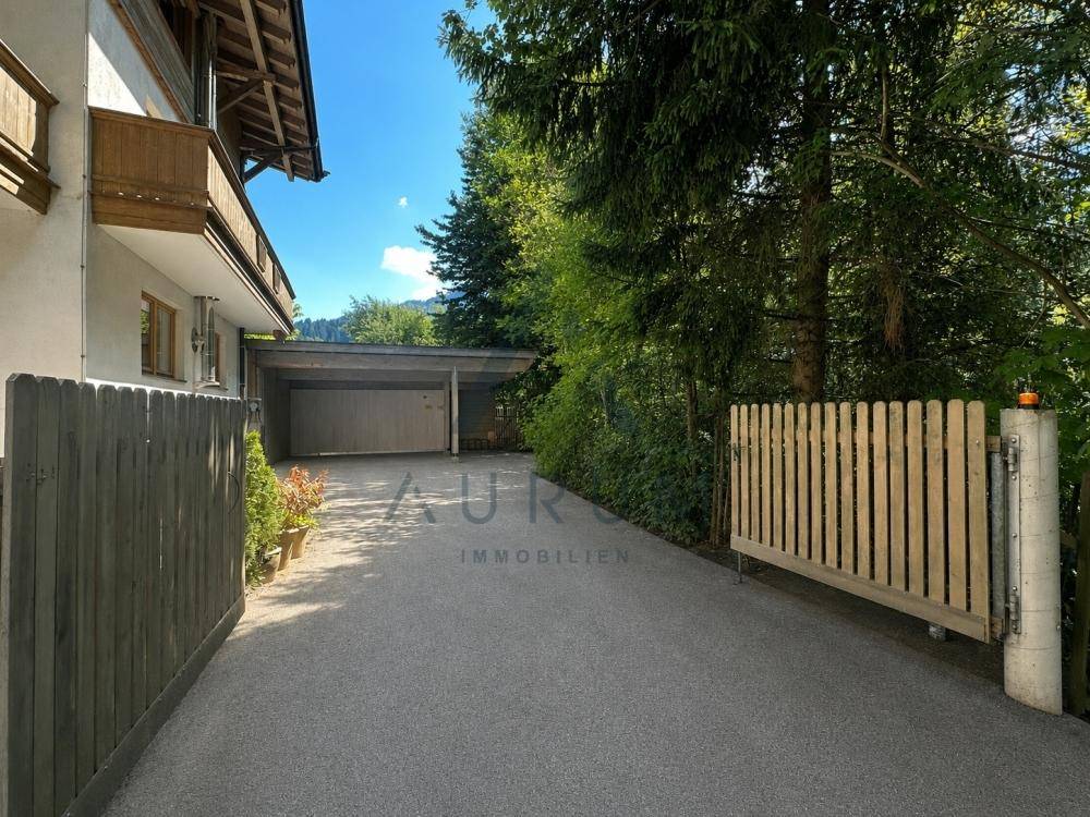 Zufahrt zum Anwesen mit Carport und umgeben von hohen Bäumen und einem Holzzaun.