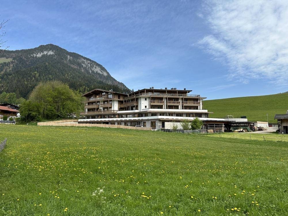 Modernes Mehrfamilienhaus in alpiner Umgebung, umgeben von grünen Wiesen und Bergen unter blauem Himmel.