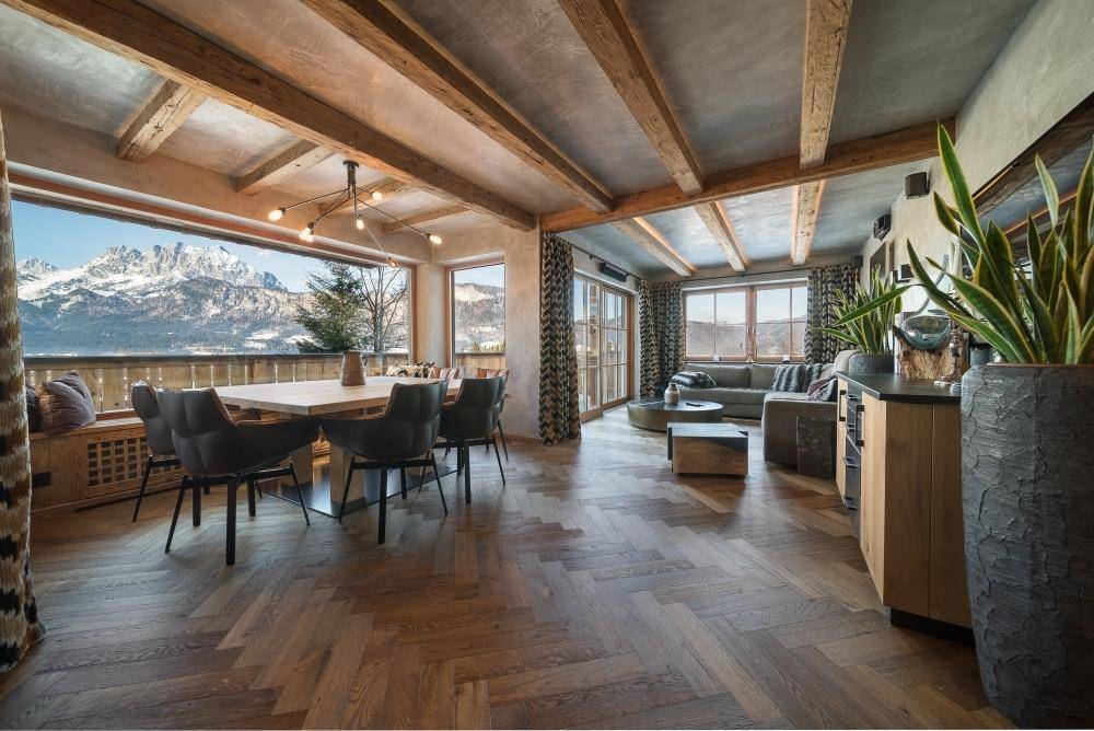 Offener Wohn- und Essbereich mit Fischgrätparkett, Holzbalken und Panoramafenstern mit Bergblick.
