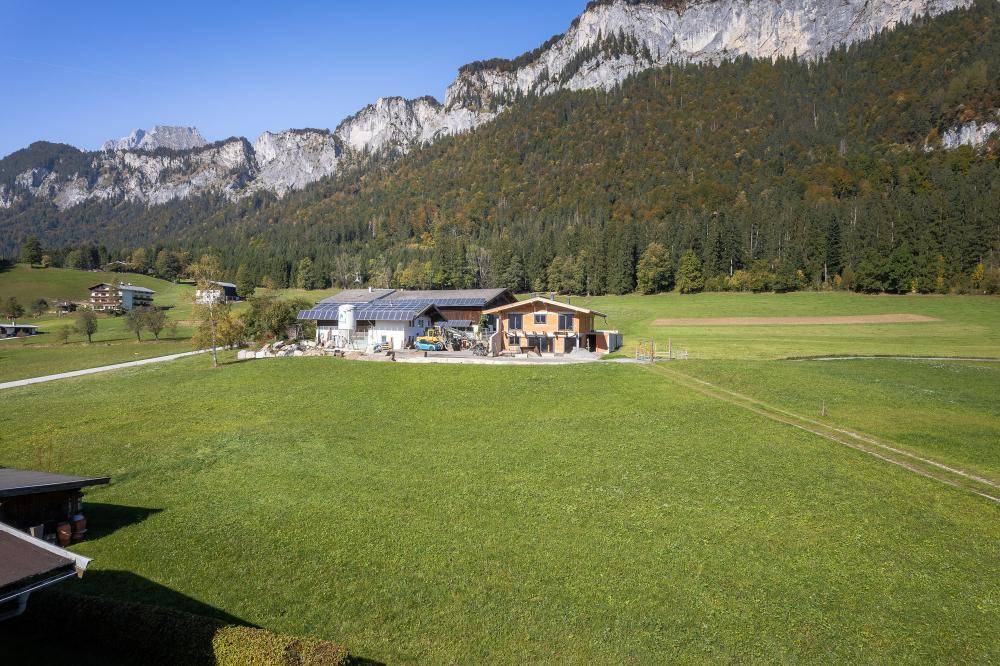 Weite grüne Wiesenfläche mit vereinzelten Häusern im Hintergrund, umrahmt von Bergen und Wald.