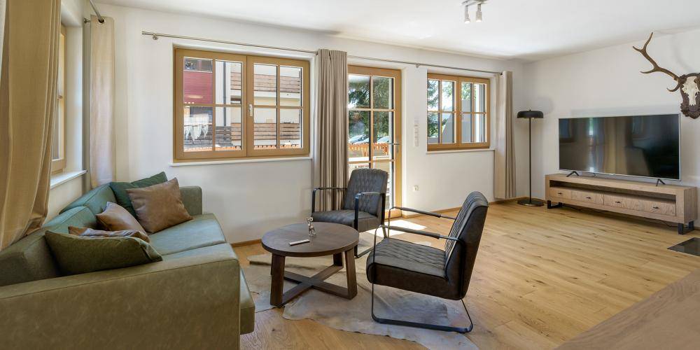 Gemütliches Wohnzimmer mit bequemer Couch, Holzboden und Blick ins Grüne durch die Fenster.