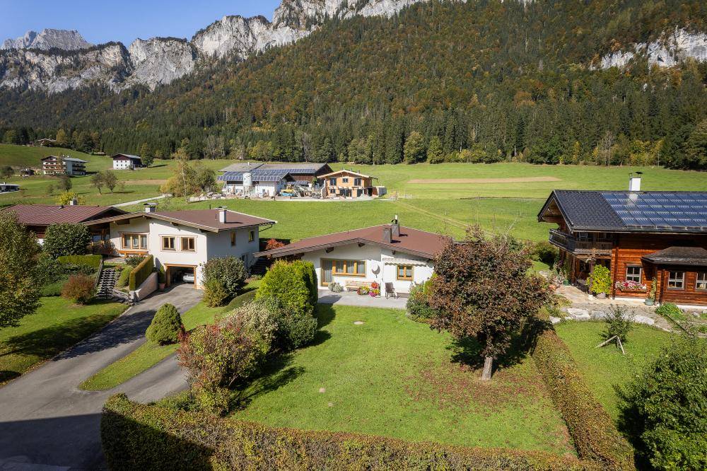 Luftaufnahme eines Hauses mit gepflegtem Garten, umgeben von grünen Wiesen und alpiner Berglandschaft.