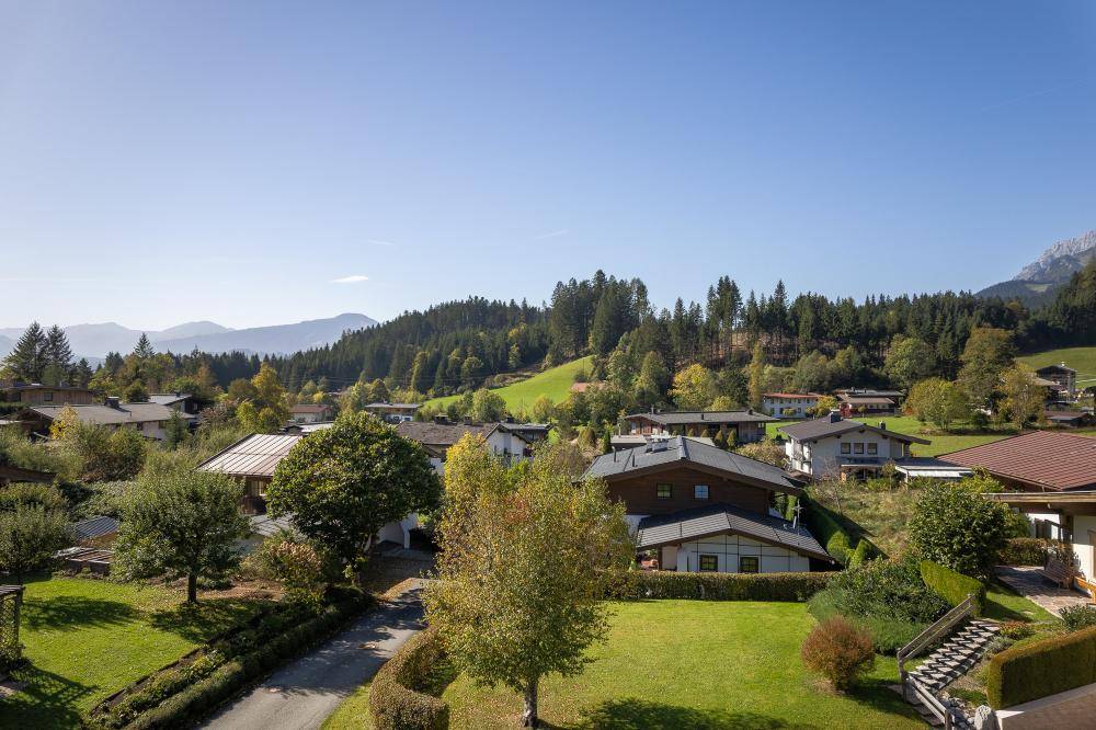 Luftaufnahme einer Wohnsiedlung mit vielen Häusern und Gärten, umgeben von Wäldern und Bergen.