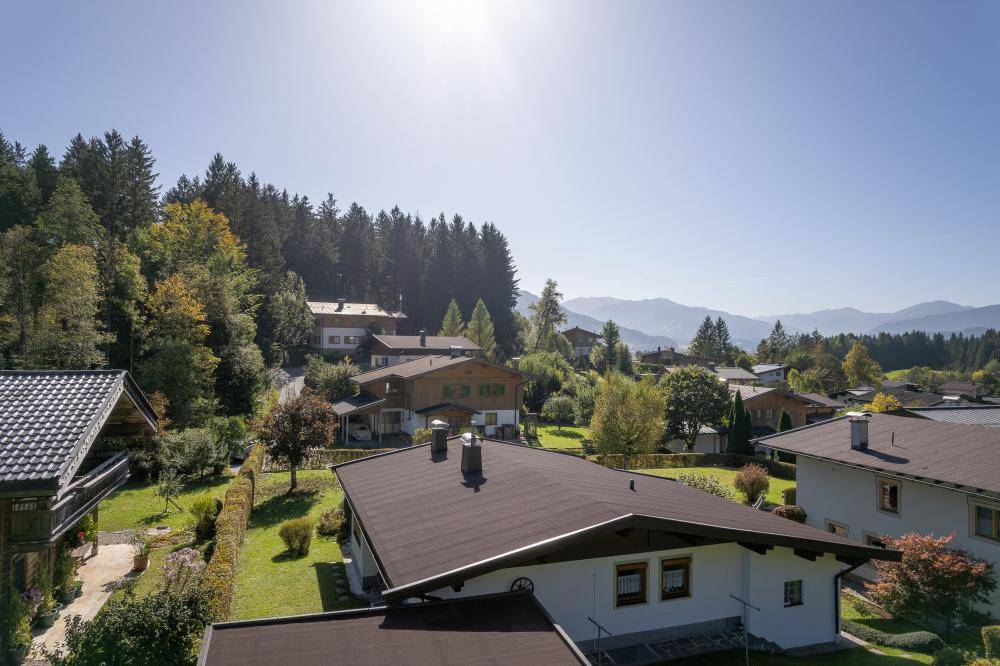 Sonnenbeschienene Luftaufnahme eines Hauses in einer Siedlung, umgeben von Wäldern und fernen Bergen.