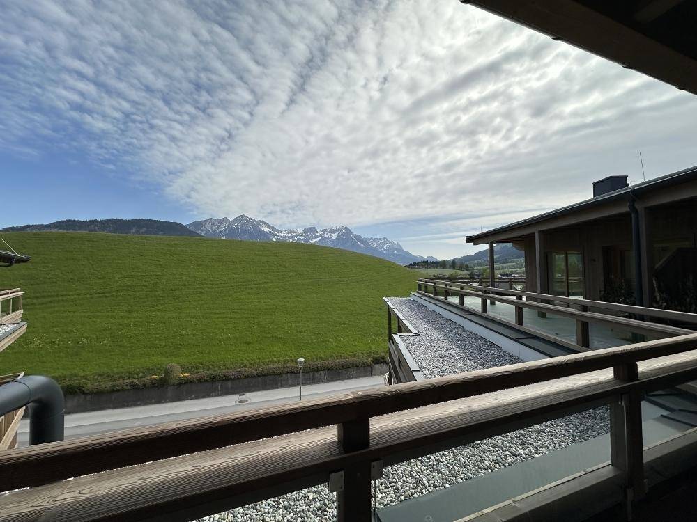 Blick von einem Balkon auf eine weite grüne Wiese und schneebedeckte Berggipfel in der Ferne.