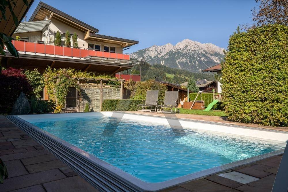 Großer Außenpool im Garten mit Liegestühlen, umgeben von Grünflächen und einem Spielplatz mit Bergblick.