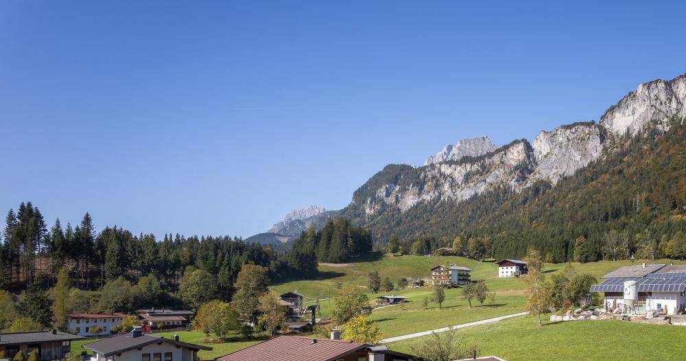 Weitläufige Landschaftsaufnahme mit majestätischen Bergen, Wäldern und vereinzelten Häusern unter blauem Himmel.