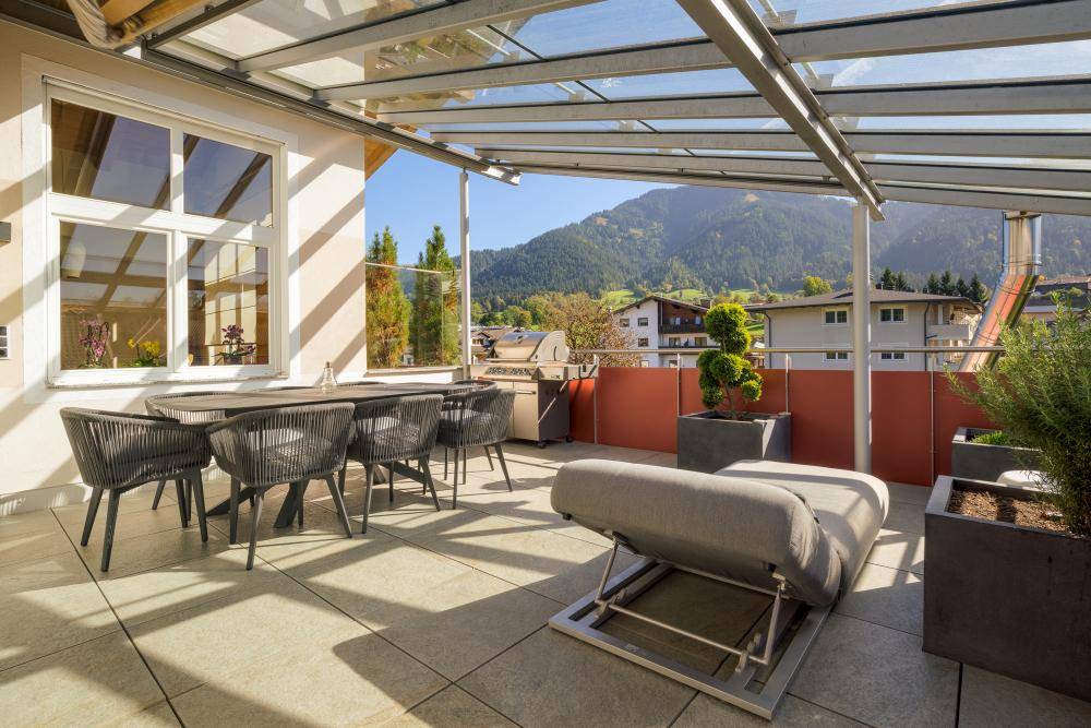 Geräumige Terrasse mit Glasdach, Essbereich und Liegefläche, umgeben von einer malerischen Berglandschaft.