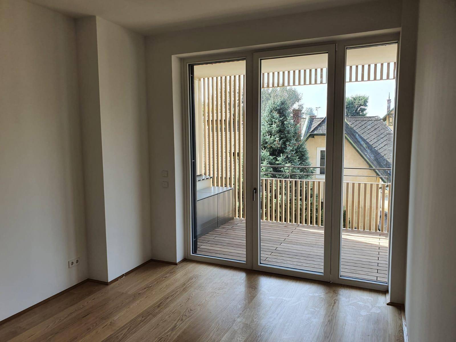Helles Zimmer mit Holzboden und großzügigem Balkonzugang, bietet einen grünen Ausblick.