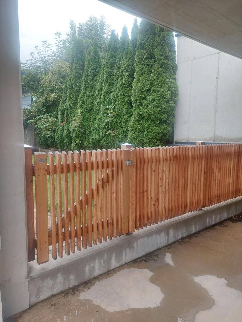 Holzzaun als Grundstücksbegrenzung mit hohen, grünen Bäumen im Hintergrund.