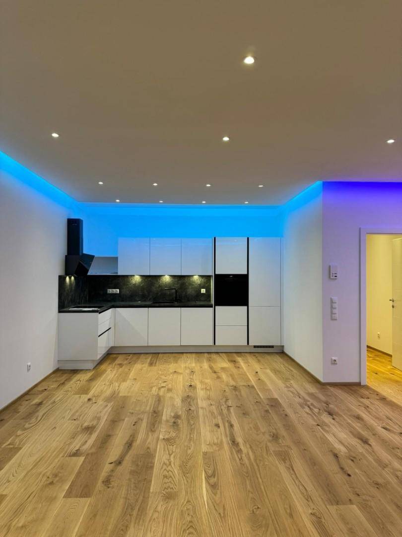 Offener Wohnbereich mit moderner weißer Küche, Holzboden und blauer LED-Akzentbeleuchtung an der Decke.