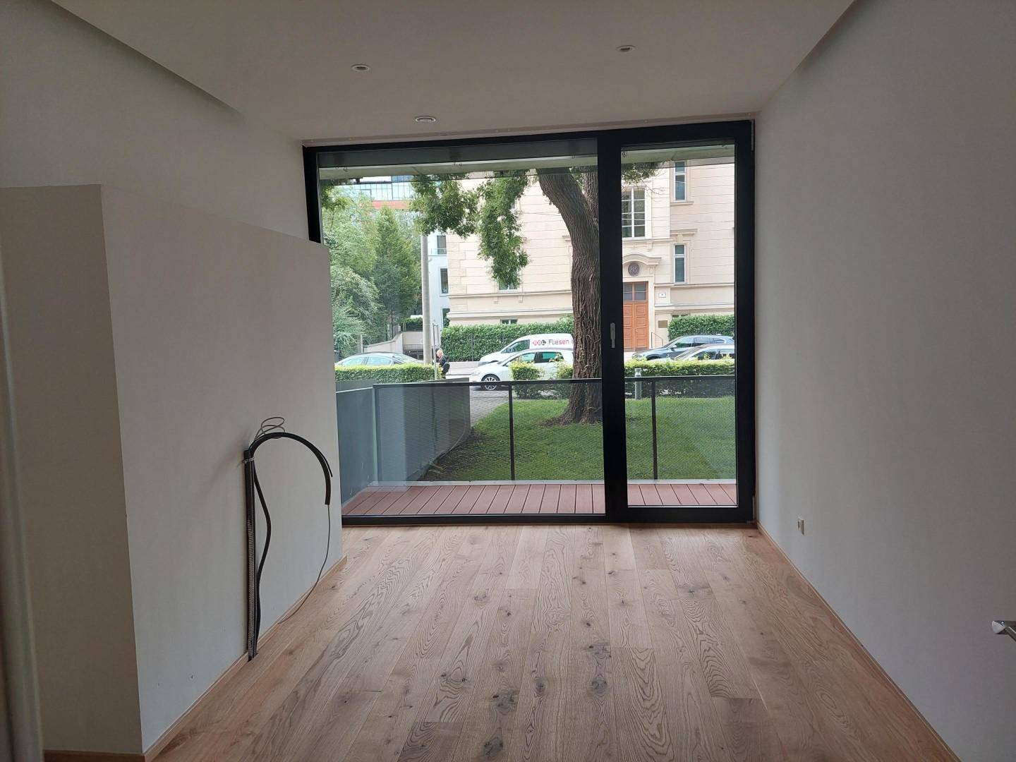 Heller Wohnraum mit Holzboden und großen Fenstern, die einen Blick auf Bäume und die Straße bieten.