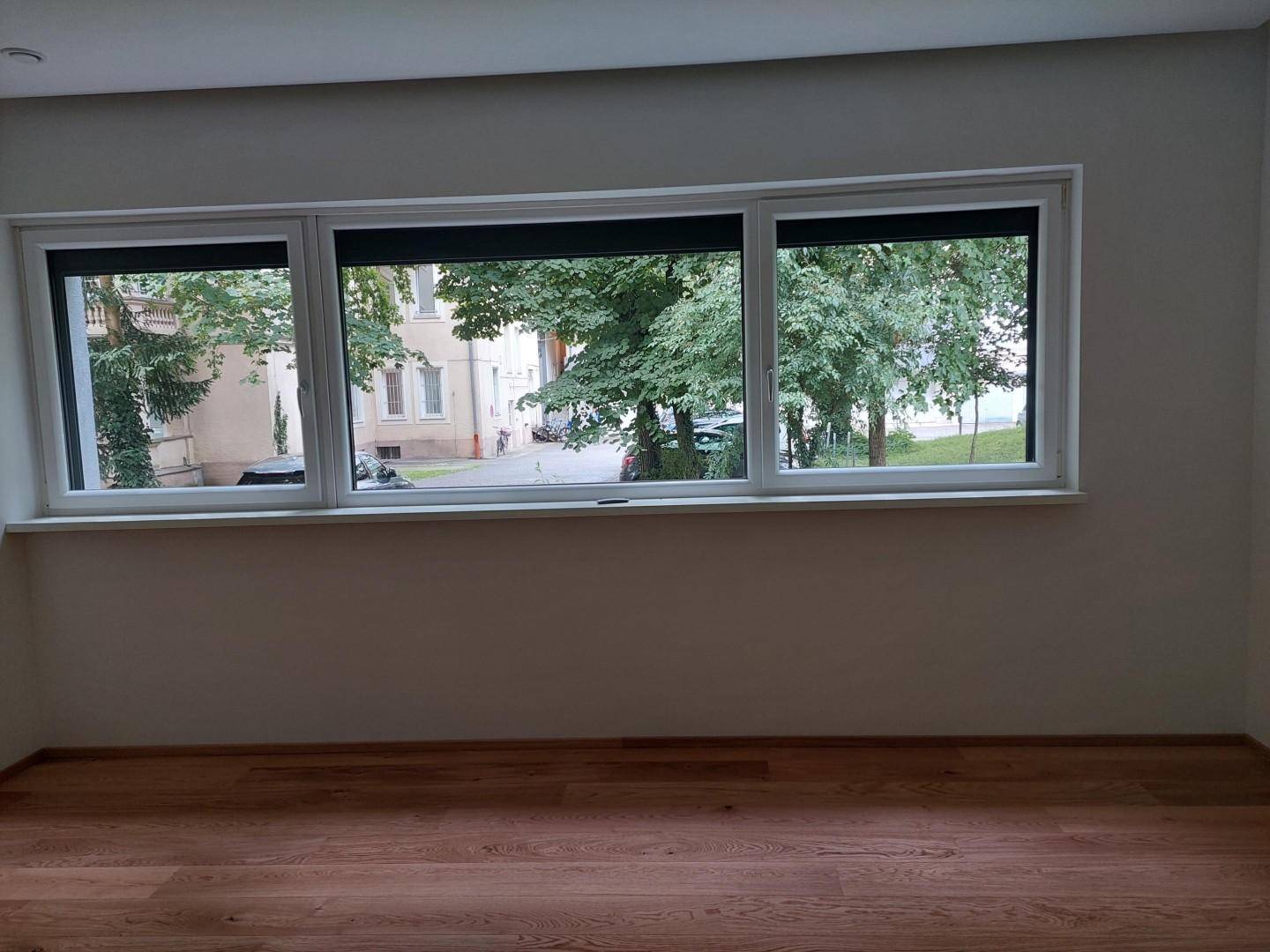 Heller Raum mit Parkettboden und Fenstern, die einen Blick auf die Straße und Bäume bieten.