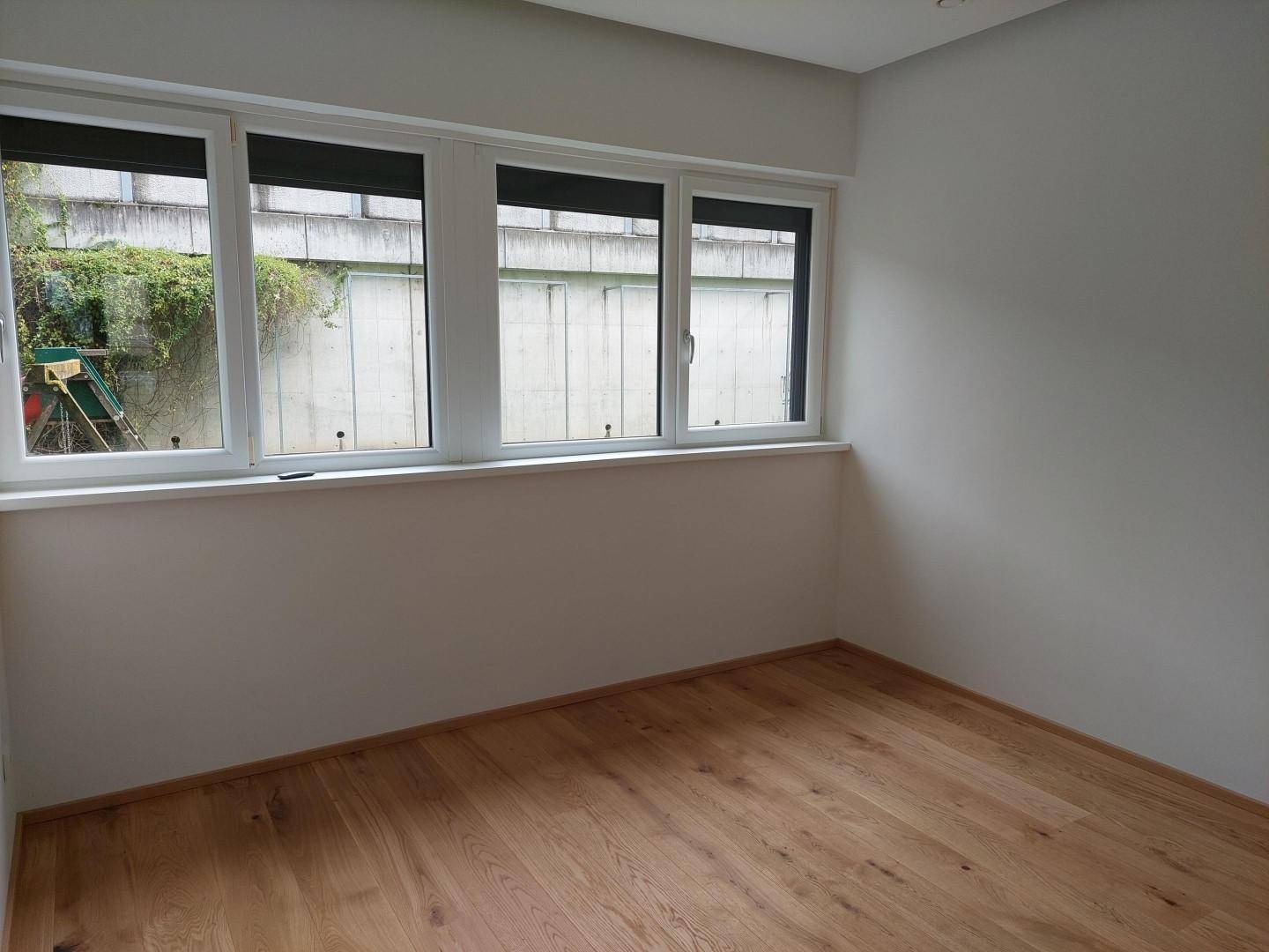 Heller, leerer Raum mit Holzfußboden und Fenstern, die eine begrünte Wand zeigen.