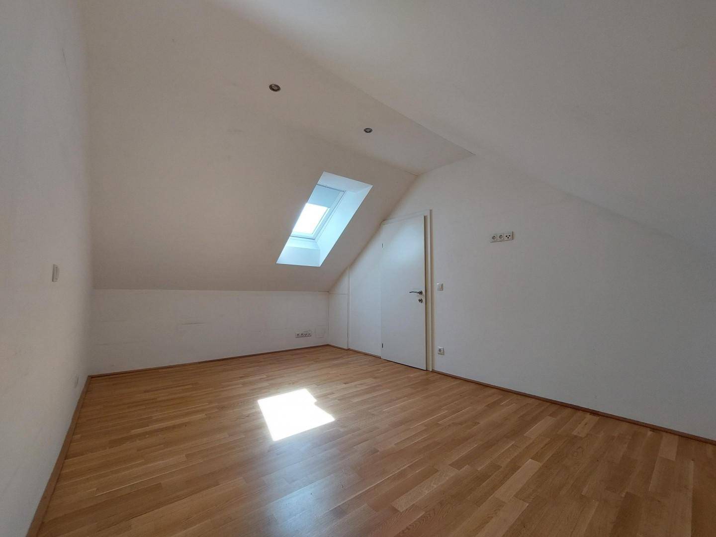 Dachgeschosszimmer mit Parkettboden, Tür und einem Dachfenster, das viel Licht hereinlässt.