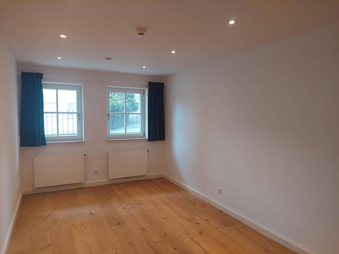 Geräumiges Zimmer mit Holzboden, zwei Fenstern und Einbaustrahlern in der Decke.