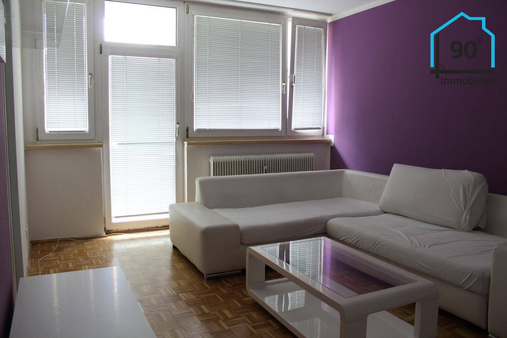 Helles Wohnzimmer mit lila Akzentwand, Parkettboden und Zugang zu einem Balkon oder einer Terrasse.