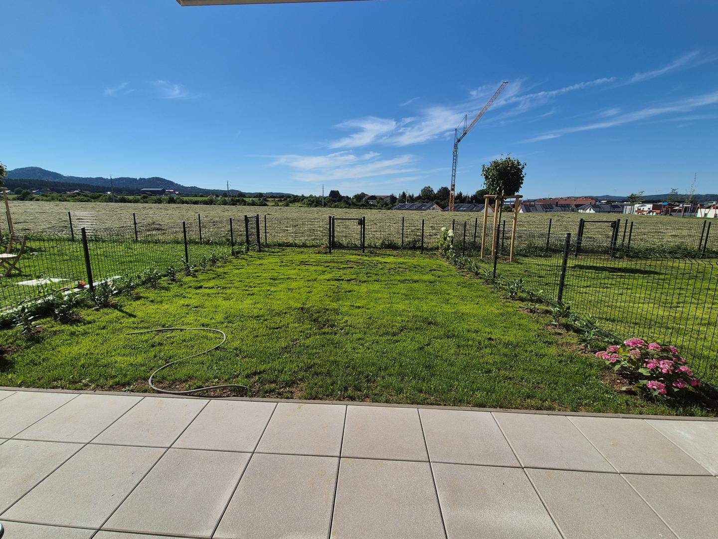 Grüner Gartenbereich mit Rasenfläche und gepflasterter Terrasse, umgeben von Feldern.