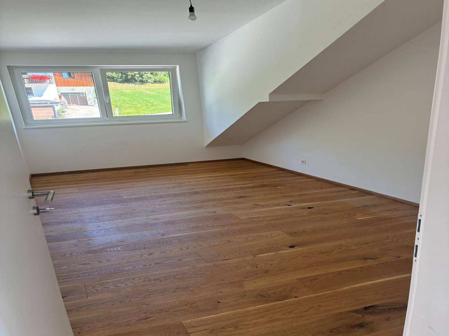 Leeres Zimmer mit Holzboden, großen Fenstern und einer Dachschräge, ideal als Schlafzimmer.