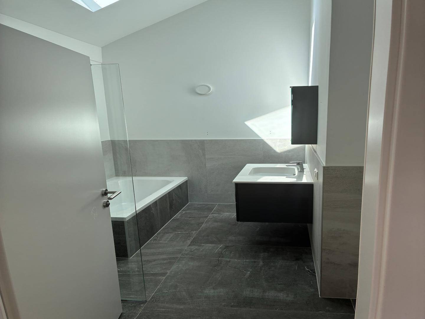 Modernes Badezimmer mit Badewanne, Waschtisch und dunklen Fliesen, beleuchtet durch ein Dachfenster.