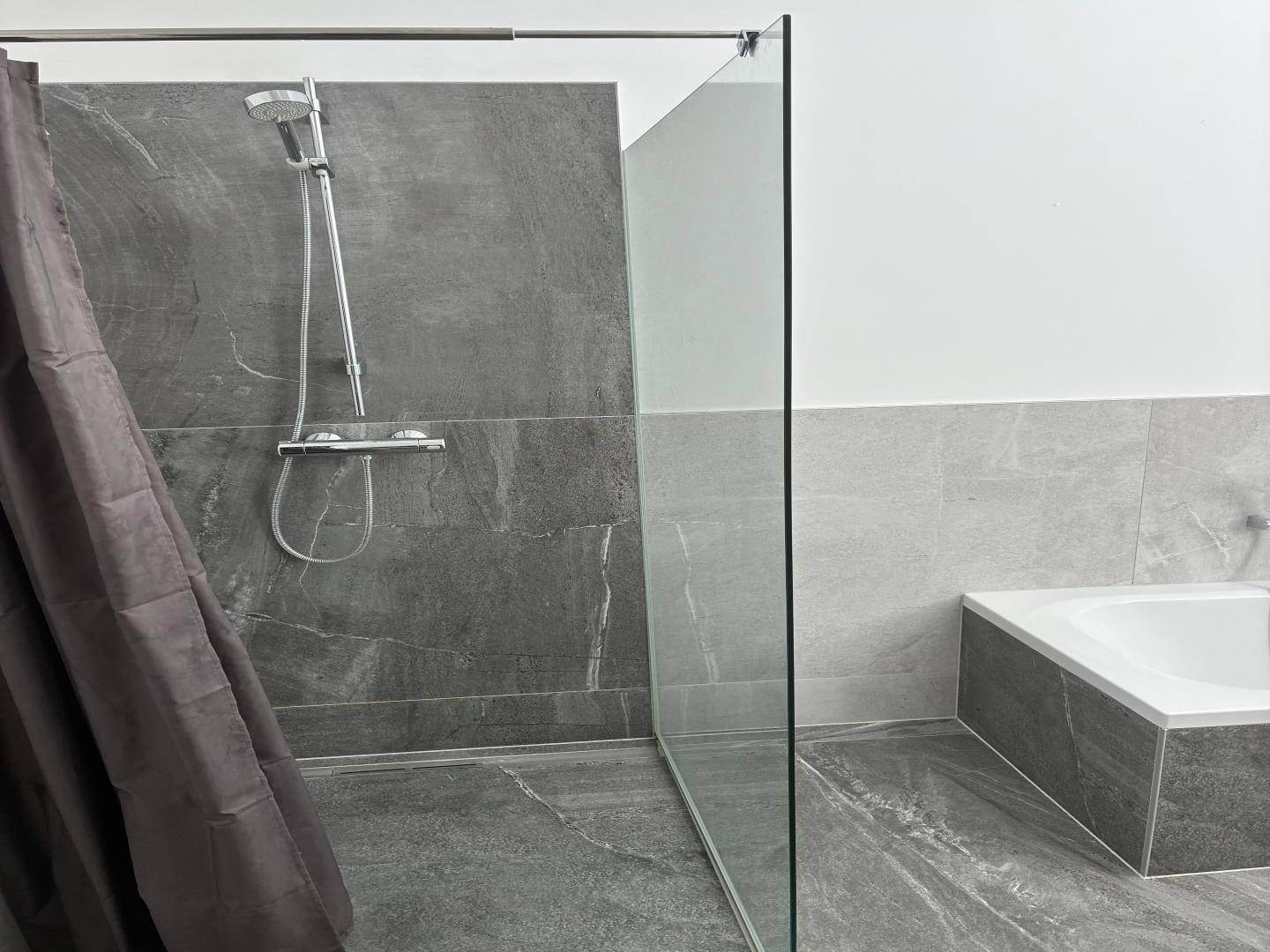 Modernes Badezimmer mit Duschbereich, Badewanne und dunklen Fliesen, durch Glaswand getrennt.