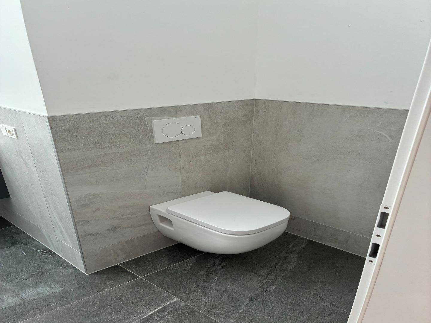 Modernes WC mit wandhängender Toilette und grauen Fliesen, sauber und funktional gestaltet.