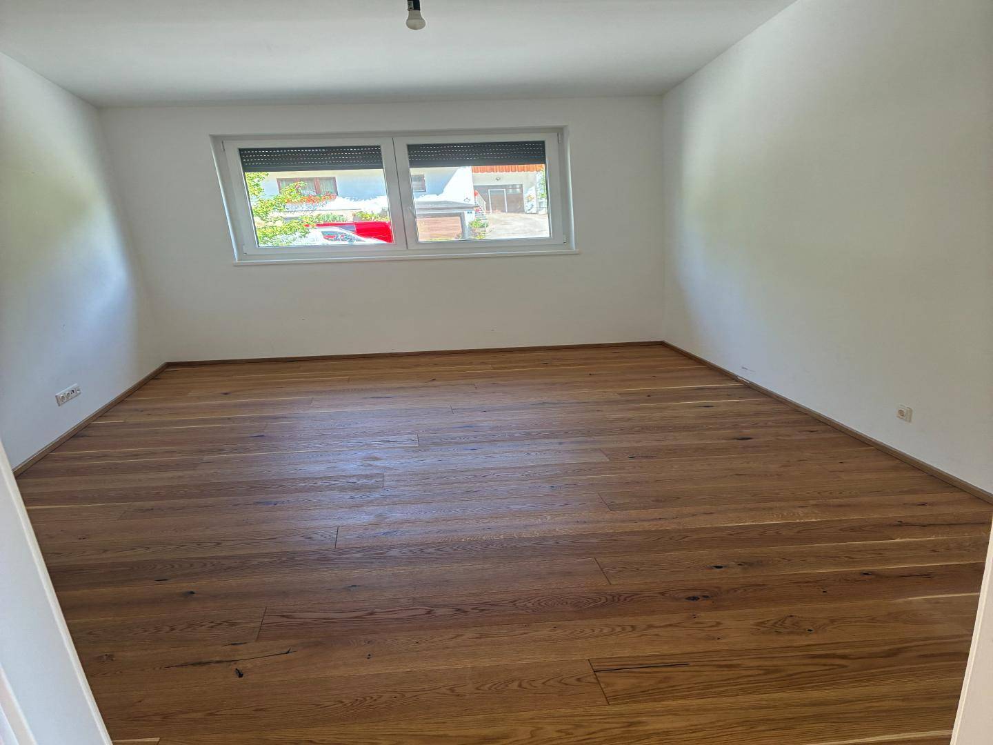 Helles Zimmer mit Holzboden und Fenster mit Rollladen, bereit zur individuellen Gestaltung.