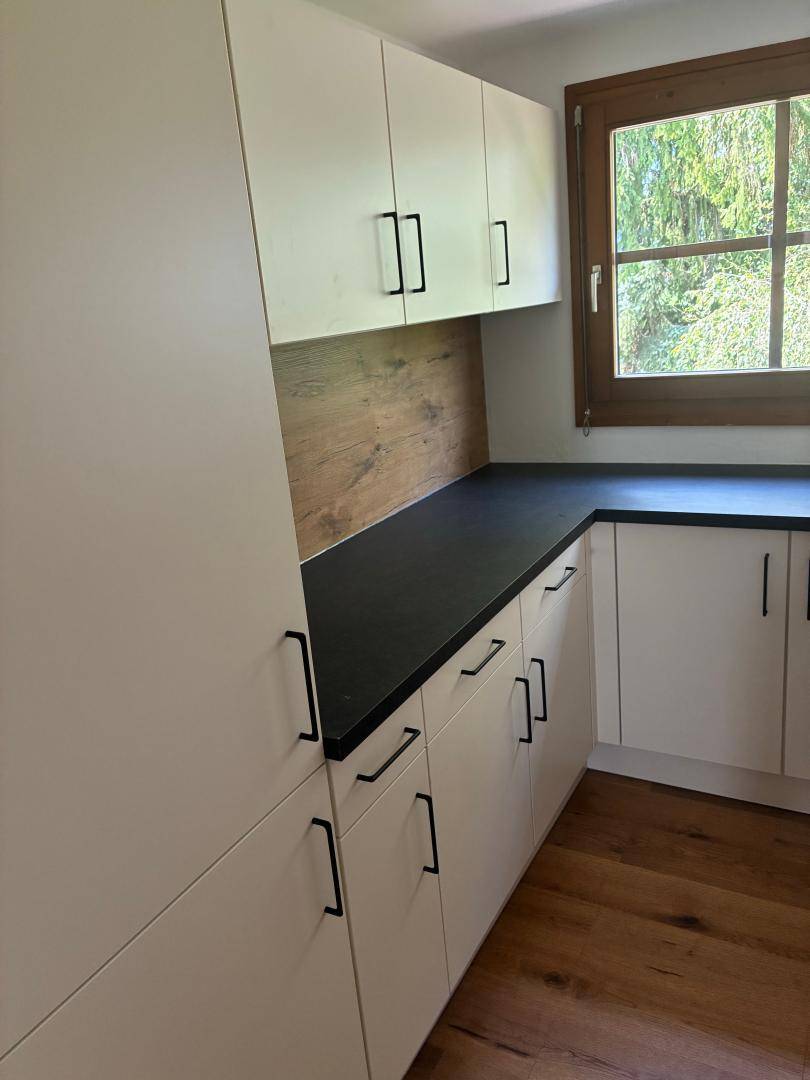 Moderne Küche mit hellen Schränken, dunkler Arbeitsplatte und Holzrückwand, bietet viel Stauraum.