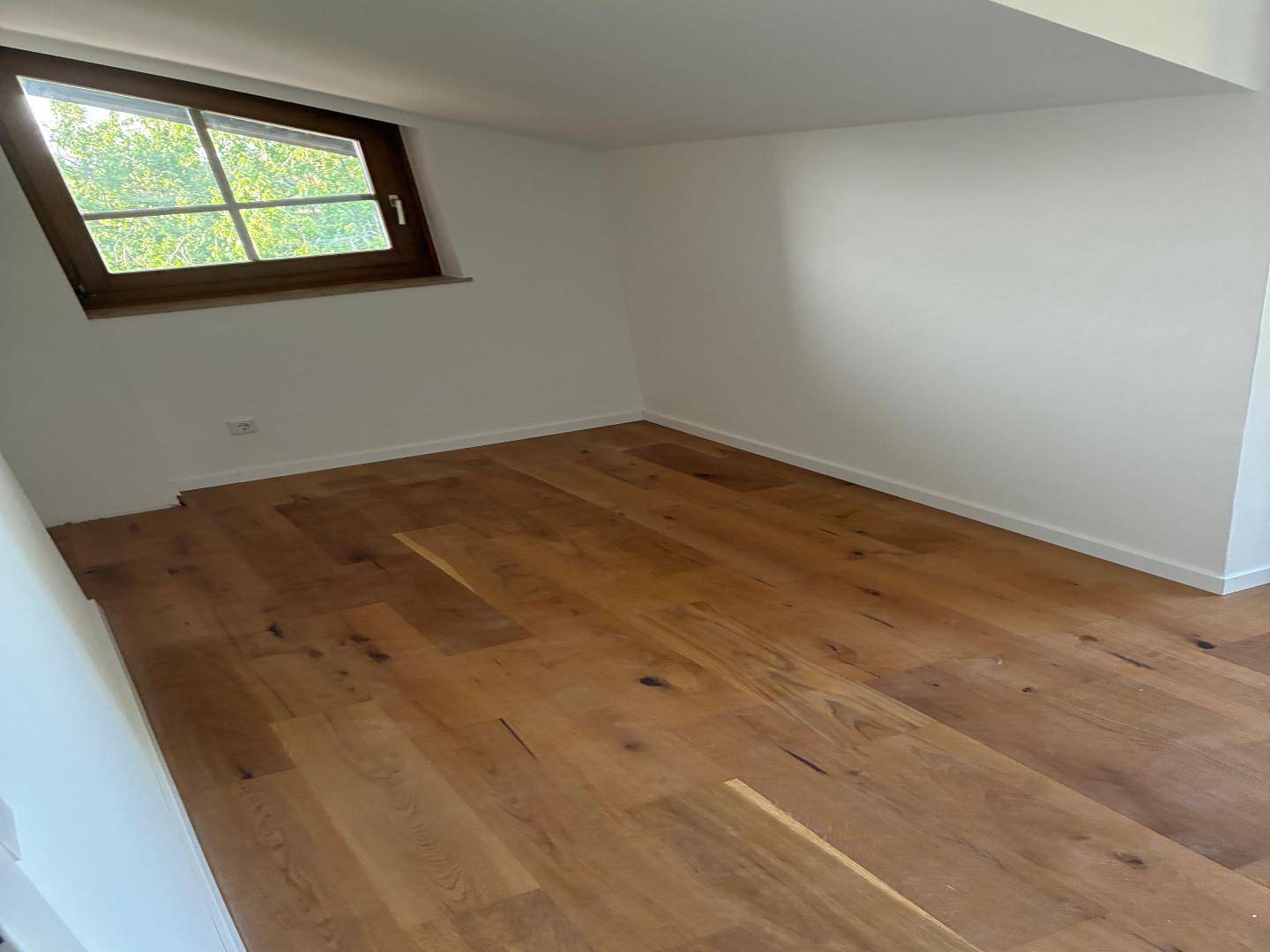 Geräumiges Zimmer im Dachgeschoss mit hellem Holzboden und viel Tageslicht durch das Fenster.